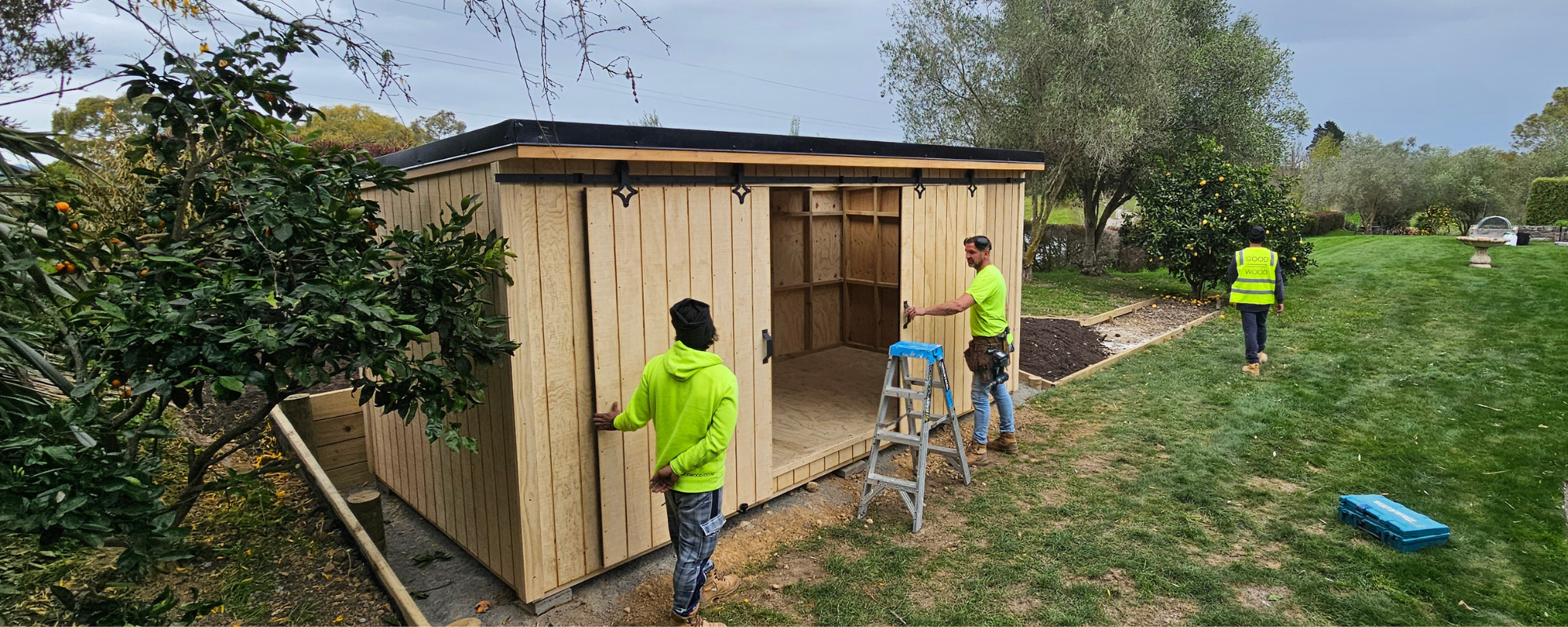 Three builders wearing hi-vis shirts working on a large wooden garden shed in a garden